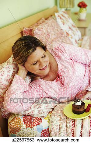 Stock Image - High angle view of a mature woman lying on the bed and holding a plate of cake. Fotosearch