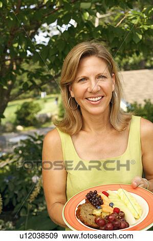 Stock Photo - Portrait of a mature woman holding a plate of fruit salad and smiling. Fotosearch