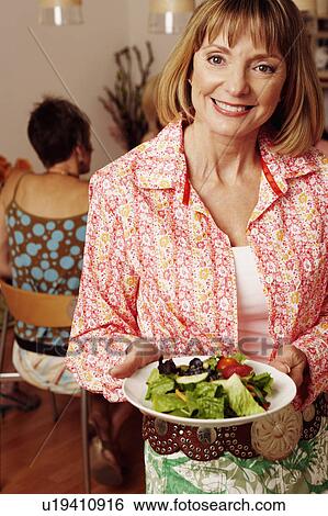 Portrait of a mature woman holding a plate of salad and her friend sitting behind her View Large Photo Image Stock Photograph - Portrait of a mature woman holding a plate of salad and her friend sitting behind her. Fotosearch