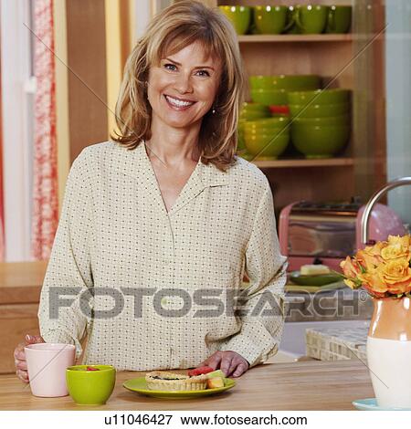Stock Photo - Portrait of a mature woman standing in the kitchen and smiling. Fotosearch