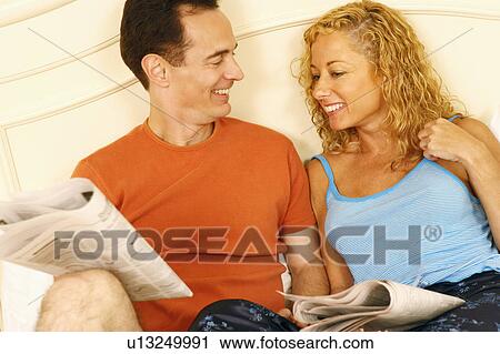 Stock Image - Close-up of a mature couple reclining on the bed and holding newspapers. Fotosearch