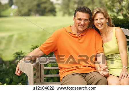 Stock Image - Portrait of a mature couple sitting on a park bench and smiling. Fotosearch