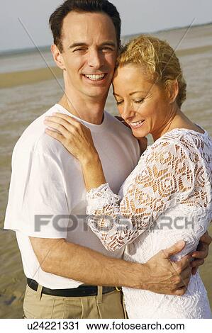 Stock Image - Side profile of a mature couple embracing each other on the beach. Fotosearch