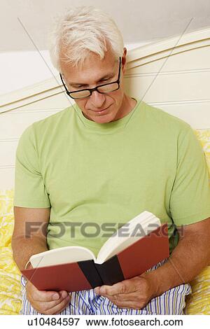 Stock Photo - Close-up of a mature man sitting on the bed and reading a book. Fotosearch