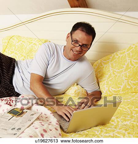 Stock Image - Portrait of a mature man lying on the bed and using a laptop. Fotosearch
