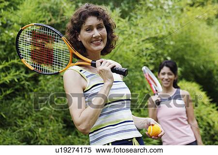 Stock Photography - Portrait of a mature woman holding a tennis ball and a tennis racket with another mature woman standing behind her. Fotosearch