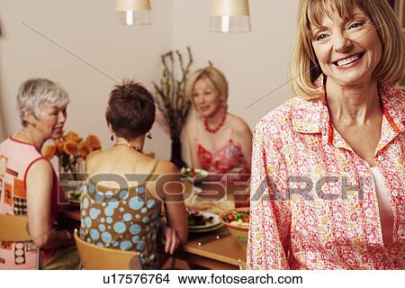 Picture - Close-up of a mature woman smiling and her friends sitting at the dining table in the background. Fotosearch