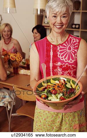 Stock Photography - Portrait of a mature woman holding a bowl of salad and her friends sitting at the dining table in the background. Fotosearch