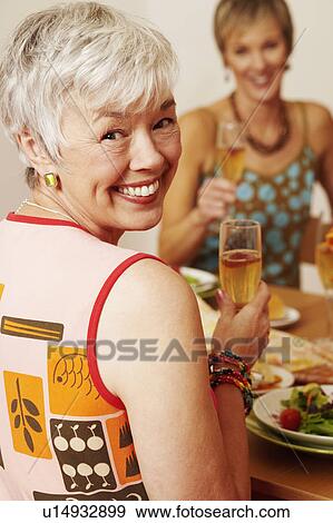 Portrait of a mature woman sitting at the dining table with her friend and holding a champagne flute View Large Photo Image Stock Photo - Portrait of a mature woman sitting at the dining table with her friend and holding a champagne flute. Fotosearch