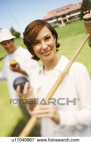 Stock Photo - Portrait of a mid adult woman holding a croquet mallet and a ball with a mid adult man in the background. Fotosearch