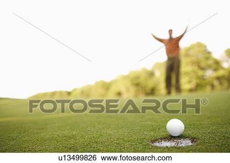 Stock Photograph - Close-up of a golf ball on the edge of a hole with a man raising his arms in the background. Fotosearch