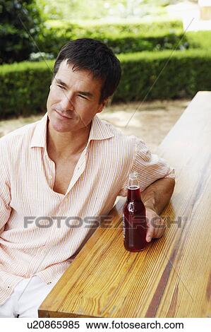Stock Photography - Close-up of a mature man sitting at a table and holding bottle of red wine. Fotosearch