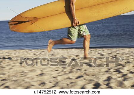 Stock Image - Low section view of a mature man holding a surfboard and walking on the beach. Fotosearch