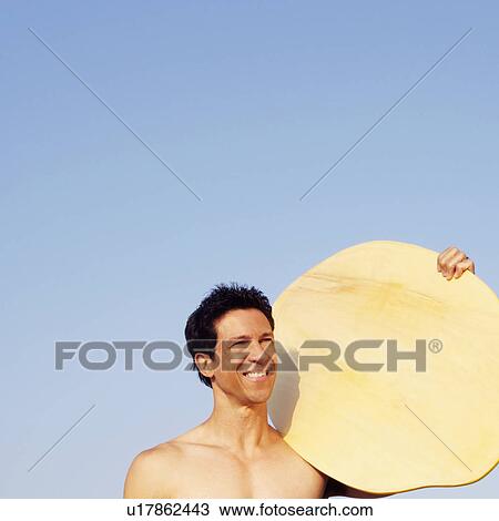 Stock Image - Mature man on the beach and holding a wooden surfboard. Fotosearch