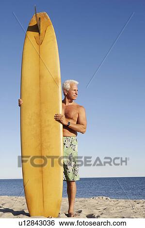 Stock Photograph - Mature man standing on the beach holding a surfboard. Fotosearch