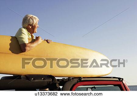 Stock Image - Side profile of a mature man sitting in a sports utility vehicle and holding a surfboard. Fotosearch