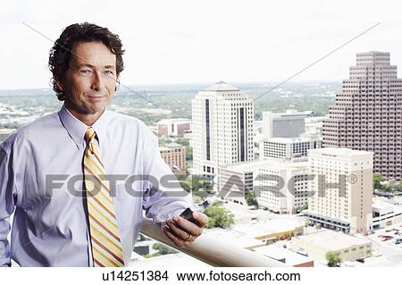 Picture - Portrait of a businessman holding a mobile phone with a city in the background. Fotosearch