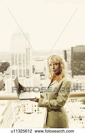 Stock Image - Portrait of a businesswoman holding a laptop with a cityscape in the background. Fotosearch