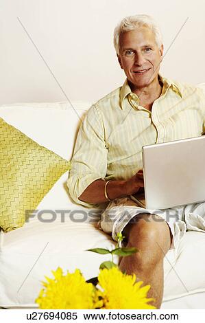 Stock Photography - Portrait of a mature man sitting on a couch and using a laptop. Fotosearch