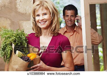 Picture - Portrait of a mature woman holding a shopping bag with a mature man talking on a mobile phone behind him. Fotosearch