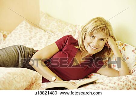 Stock Image - Portrait of a mature woman lying on the bed and reading a book. Fotosearch