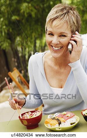 Picture - Close-up of a mature woman eating with chopsticks and talking on a mobile phone. Fotosearch