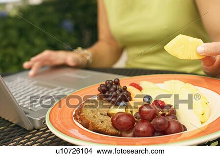 Mid section view of a woman using a laptop and eating fruit salad View Large Photo Image Picture - Mid section view of a woman using a laptop and eating fruit salad. Fotosearch