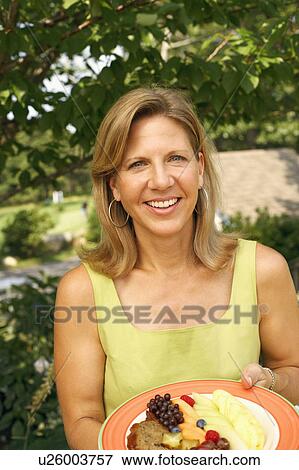 Stock Photo - Portrait of a mature woman holding a plate of fruit salad and smiling. Fotosearch