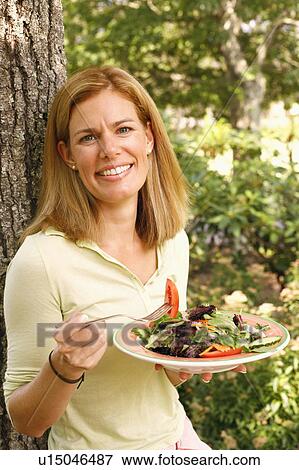 Stock Photo - Portrait of a mid adult woman leaning against a tree and eating a vegetable salad. Fotosearch