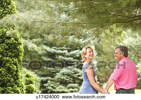 Stock Image - Mature couple holding hands and smiling in a park. Fotosearch