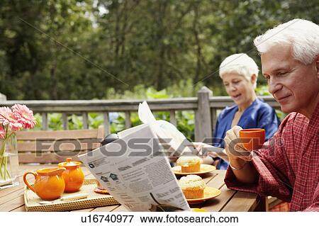 Stock Photo - Close-up of a mature man holding a cup and reading a newspaper with a senior woman in the background. Fotosearch