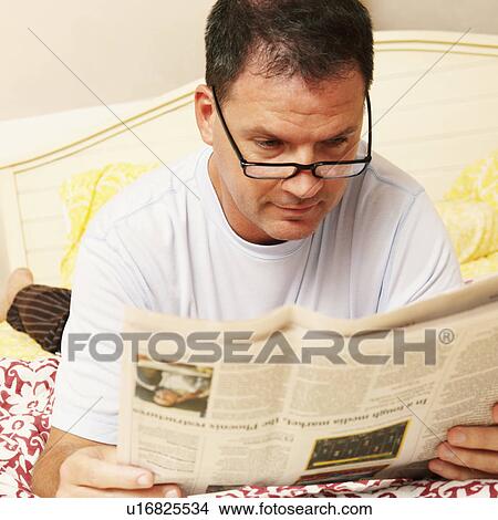 Picture - Close-up of a mature man lying in bed and reading a newspaper. Fotosearch