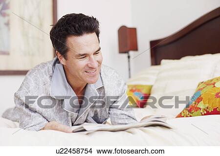Picture - Mature man lying on the bed reading a newspaper. Fotosearch