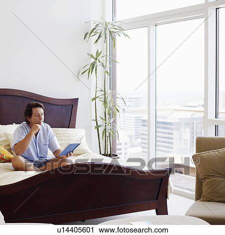 Stock Image - Mature man sitting on the bed reading a book. Fotosearch