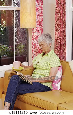 Stock Image - Senior woman sitting on a couch and reading a book. Fotosearch