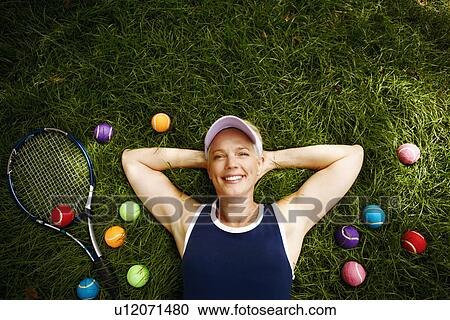 Stock Image - High angle view of a mature woman lying on the grass with tennis balls and a tennis racket. Fotosearch