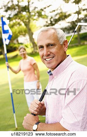 Stock Image - Portrait of a mature man holding a golf club with a mature woman holding a golf flag in the background. Fotosearch