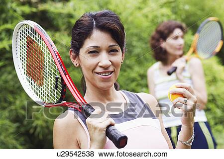 Stock Photo - Portrait of a mature woman holding a tennis ball and a tennis racket with another mature woman standing behind her. Fotosearch