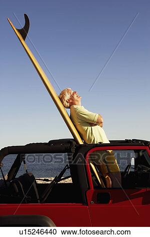 Stock Image - Side profile of a mature man leaning against a surfboard in a sports utility vehicle. Fotosearch