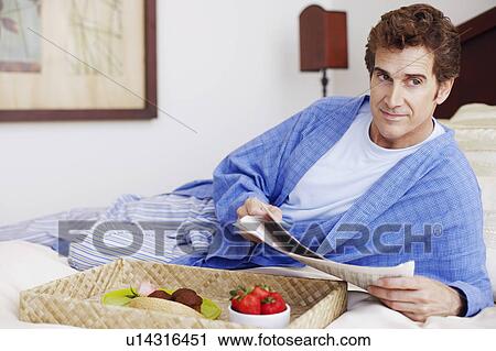 Stock Image - Close-up of a mature man lying on the bed and holding a newspaper. Fotosearch