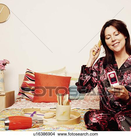 Mature woman sitting on the bed with a flip phone and combing her hair View Large Photo Image Picture - Mature woman sitting on the bed with a flip phone and combing her hair. Fotosearch