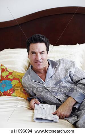 Picture - Portrait of a mature man lying on the bed holding a newspaper. Fotosearch