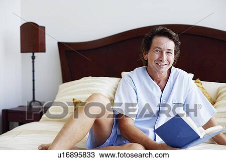 Stock Image - Portrait of a mature man sitting on the bed with a book and smiling. Fotosearch
