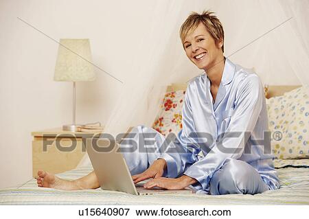 Stock Photo - Portrait of a mature woman using a laptop on the bed and smiling. Fotosearch