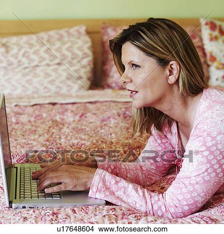 Picture - Side profile of a mature woman lying on the bed and using a laptop. Fotosearch