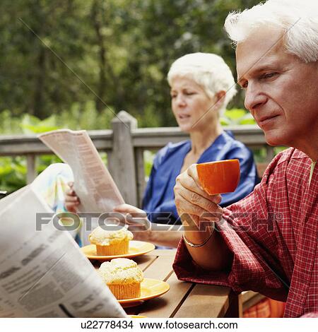 Picture - Close-up of a mature man holding a cup and reading a newspaper with a senior woman in the background. Fotosearch