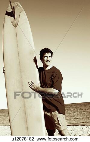 Stock Image - Mature man standing on the beach holding a surfboard. Fotosearch