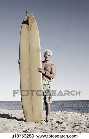 Stock Photo - Portrait of a mature man standing on the beach holding a surfboard. Fotosearch