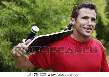 Stock Image - Portrait of a mature man carrying a skateboard on his shoulder and smiling. Fotosearch