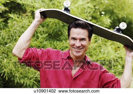Stock Image - Portrait of a mature man holding a skateboard on his head and smiling. Fotosearch